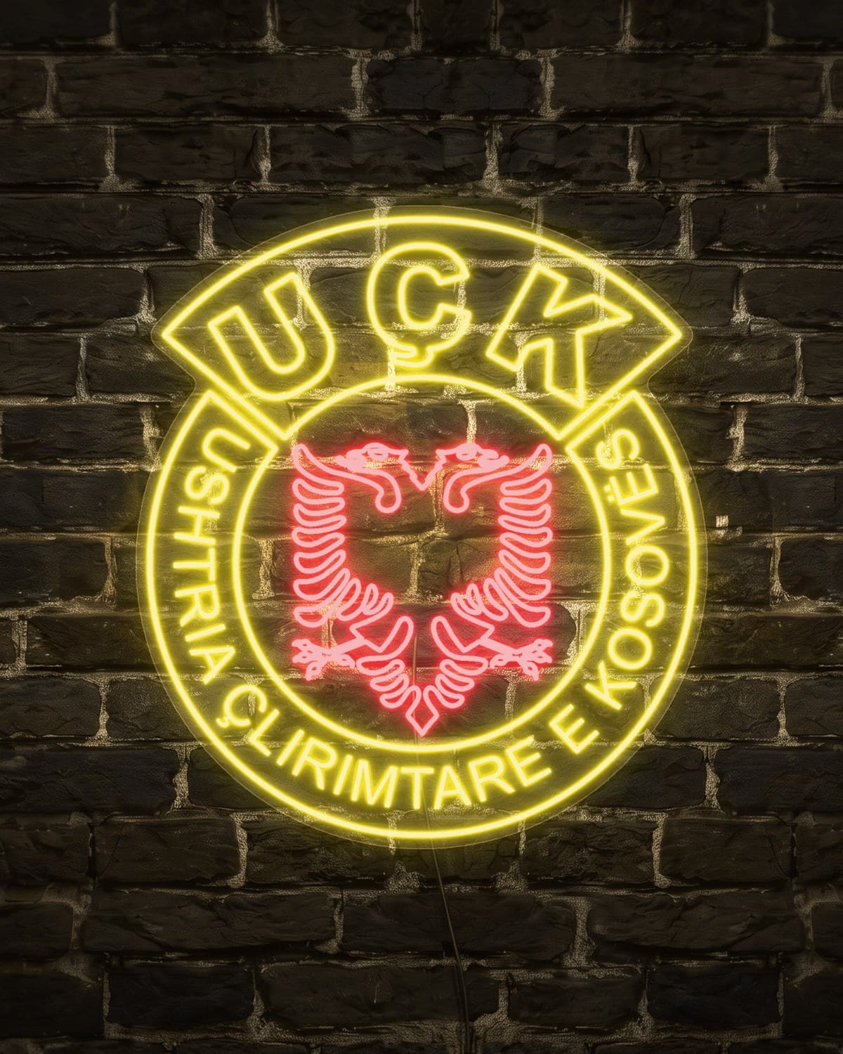 Emblema e UÇK-së si tabelë neon LED e verdhë dhe e kuqe, e ndezur në mur me tulla